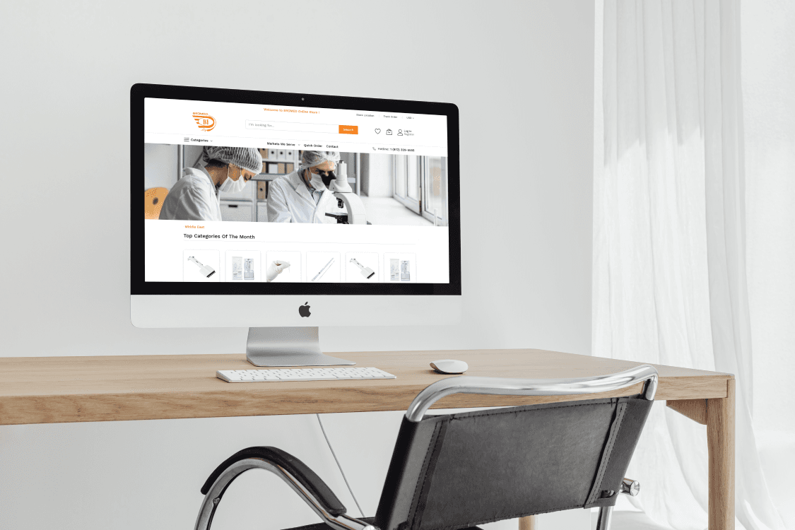 An Apple iMac on a wooden desk displaying the homepage of the BROMED medical supplies website by Brosco International, showing scientists in a lab and top product categories.
