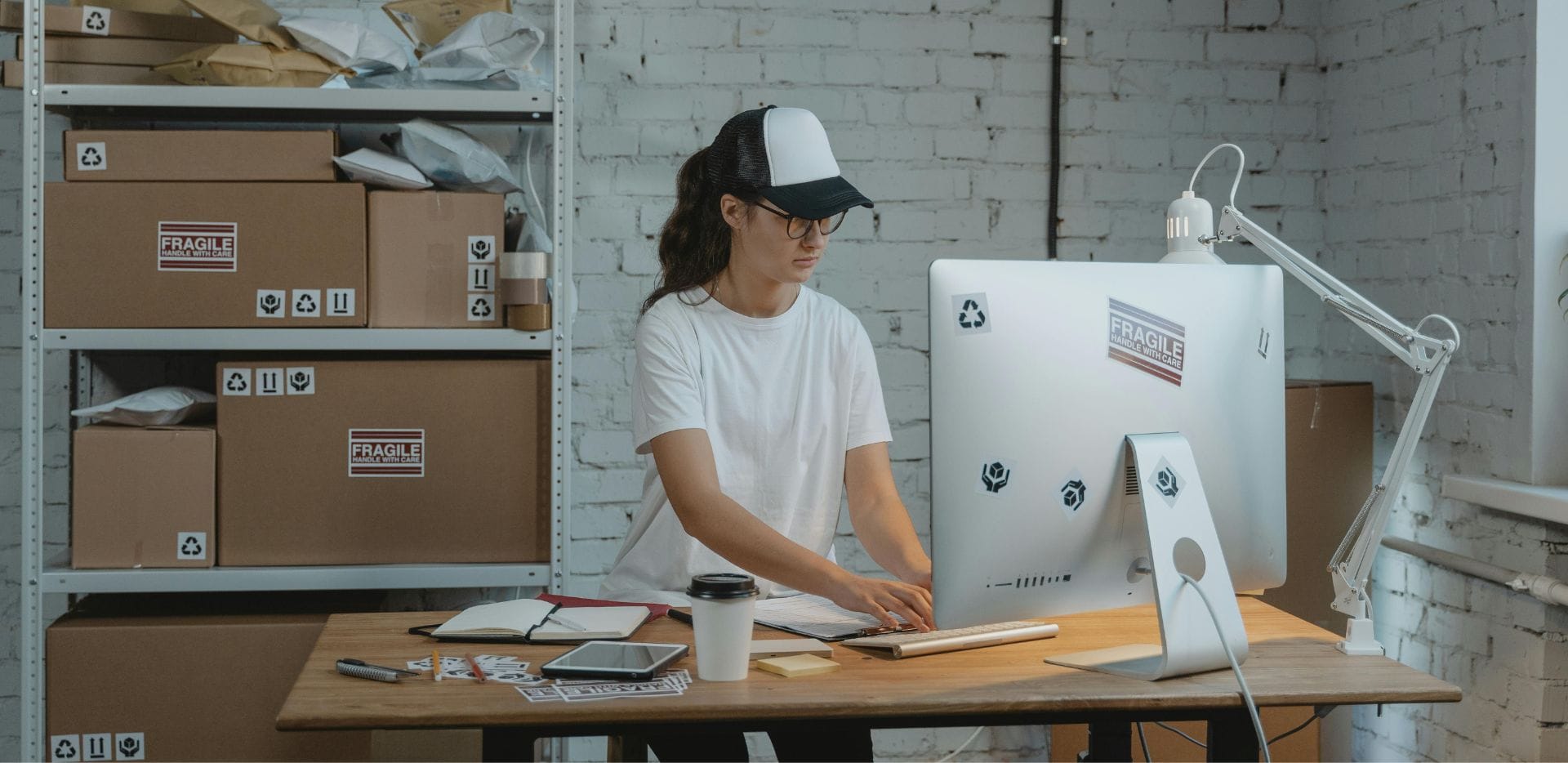 A person in a white t-shirt and cap working intently at a computer in a shipping office, surrounded by boxes and shelves, symbolizing efficient order processing and inventory management for the EGC Supply Magento case study by Ceymox.